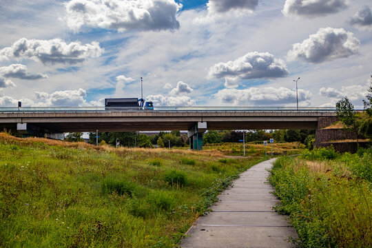 Plain with a pedestrian path, elevated motorway bridge with a truck driving in background, against blue sky and white clouds, wild grass, sunny summer day in South of Limburg, Netherlands