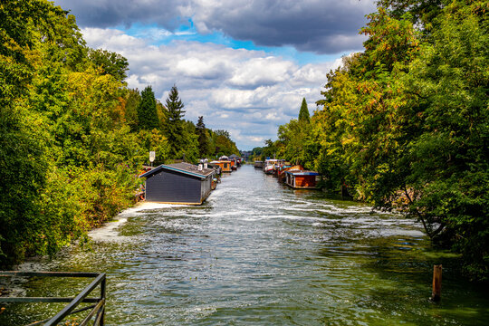 Navigation canal with floating cityscape showing houseboats on shore outside the city of Maastricht, surrounded by abundant leafy trees against a cloudy sky in background, South Limburg, Netherlands