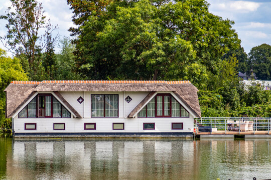 Picturesque houseboat with white walls, thatched roofs and large windows, a small terrace, leafy green trees in the background, sunny day in Maastricht, South Limburg, Netherlands