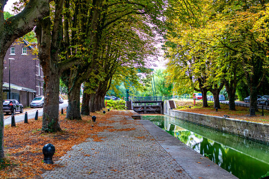 Manual lock number 19 and navigation canal for small boats, cobbled sidewalk between autumn trees, bridge with wooden gates in background, sunny day in city of Maastricht, South Limburg, Netherlands