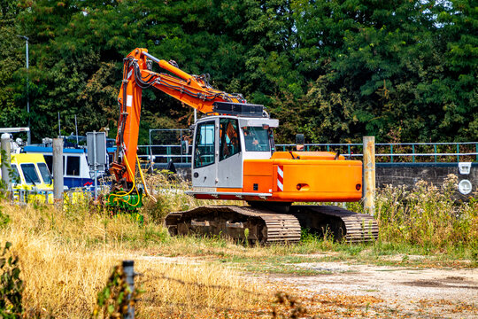 Orange tracked excavator on a plot of dry wild grass next to dock on navigable canal, partial view of boats in the background, industrial machinery, sunny day in South Limburg, Netherlands