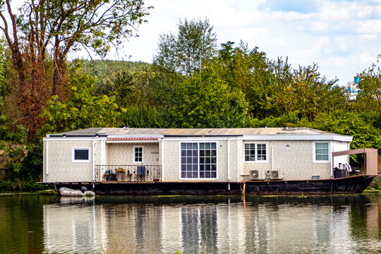Floating house with white walls on navigation canal, large windows and balcony, calm waters with reflection on water surface, leafy trees in background, cloudy day in South Limburg, Netherlands