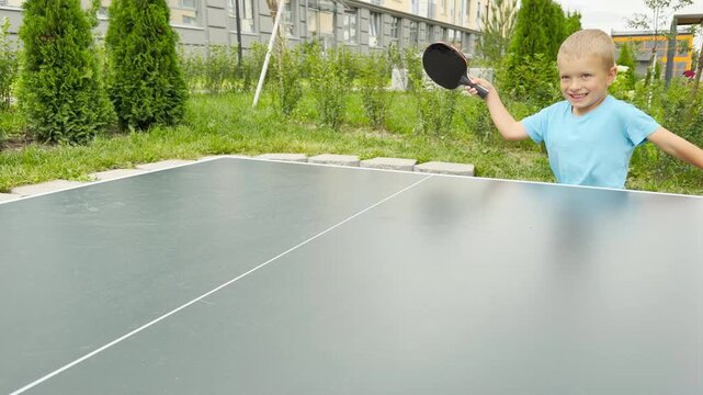 A smiling young boy holds up his table tennis paddle with cheerful enthusiasm. He stands by an outdoor ping pong table in a bright green courtyard.