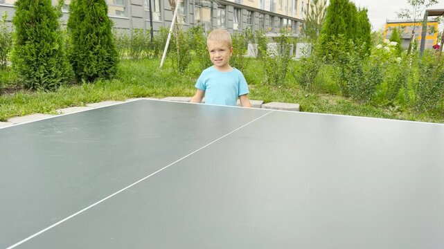 A focused little boy raises his paddle to play table tennis with joyful determination. He stands at an outdoor court surrounded by lush green bushes.