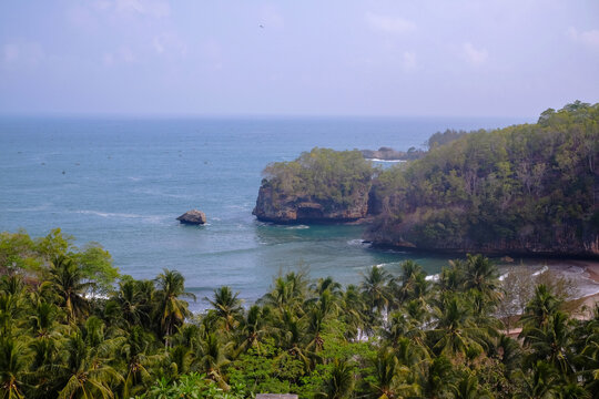 A stunning high-angle view featuring the blue sea, lush green coconut trees, and a rocky bay in Pacitan, Indonesia. Perfect for a summer travel theme.