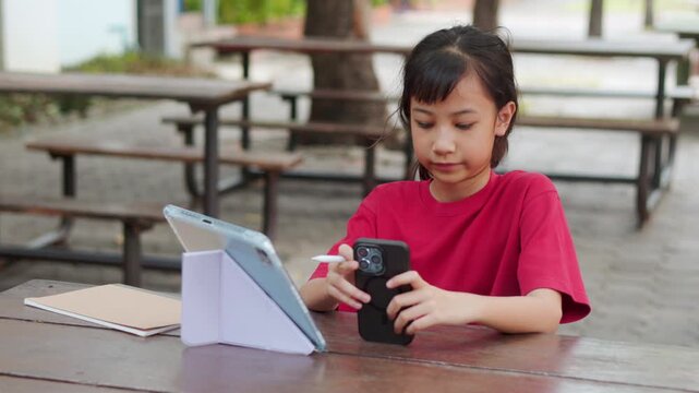 Asian kid doing homework on digital tablet at outdoor campus table. The selective focus shows the student enjoying active e-learning, highlighting modern education technology and tech-savvy childhood.