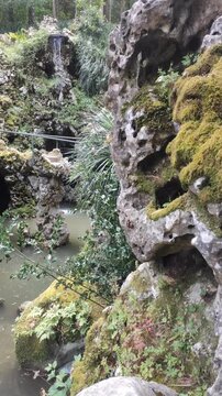 Scenic view of an artificial grotto with mossy rock formations, lush green foliage, and a small cascading waterfall dropping into a calm pond in a natural park or garden setting