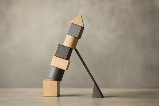 A precarious stack of wooden geometric blocks balanced on a dark support against a textured.