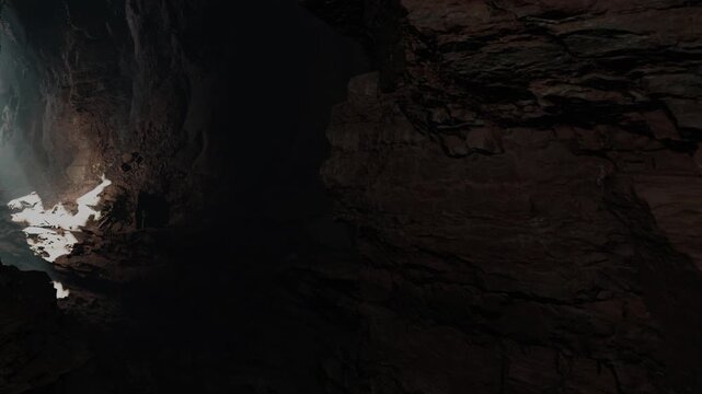 A beam of light enters a cave showing rock formations and dark areas. The interior has rough textures and shadows. The scene captures the nature of a hidden underground space.