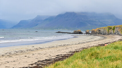 Breathtaking view of a soft white sand beach stretching along the Westfjords of Iceland, with towering mountain peaks rising into the clouds on a dramatic summer day. © Asanee