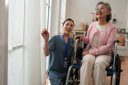 An elderly female patient with Alzheimer's disease is in a wheelchair, with an Asian nurse sitting beside her, holding her hand, offering encouragement, and pushing the wheelchair, caring for the elde
