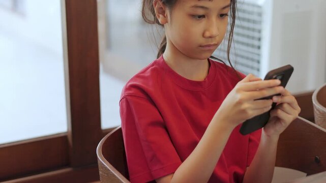 Close up Asian kid seriously focusing on a smartphone at a restaurant. Thai girl intently playing mobile games or watching content. Shot with selective focus and copy space. Digital lifestyle concept.