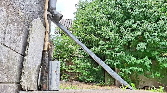 A weathered rural house wall and old metal drainpipe frame a quiet village yard, surrounded by lush trees, rustic fencing, greenery, and natural overgrowth in peaceful countryside