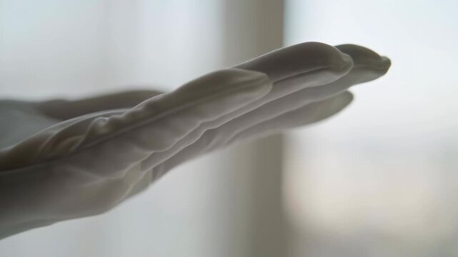 Gloved hand slowly turns with fingers extended in close-up beside a bright window, moving from palm-facing view toward a side angle under soft backlight and shallow focus