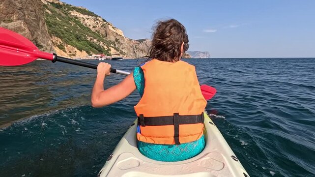 Paddling single kayak along scenic mountainous shore on calm ocean water during sunny afternoon. Rear view tracking shot.