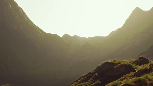The sun rises over the mountains, lighting up the peaks and creating shadows. Green plants grow at the base of the hills, showing natures beauty in a quiet morning.