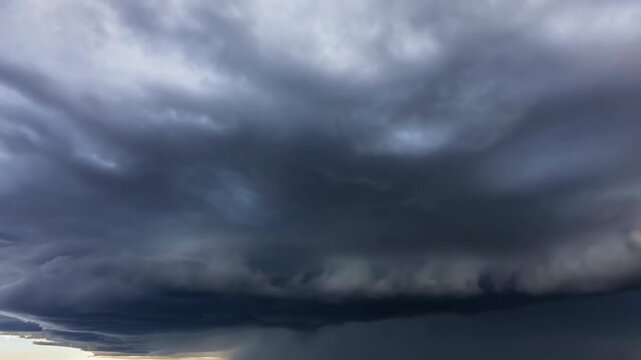 Extensive dark storm clouds roll across an open horizon, forming layered swirling bands beneath a turbulent gray sky with a narrow strip of pale light visible near the distant landscape