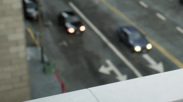 Cars drive down a city street during the day. The view is taken from a higher point, showing traffic and the street layout below. Buildings are visible in the background.