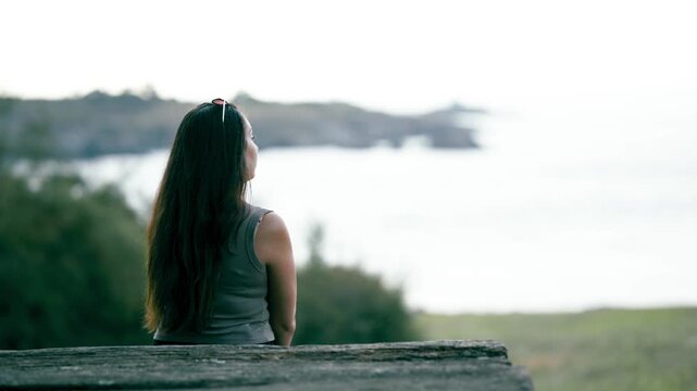 Thoughtful woman sitting alone on a rustic wooden bench. She is quietly observing the peaceful ocean and coastline from a scenic viewpoint