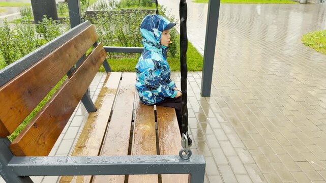 A young boy in a blue jacket sits on a wooden swing and eats candy outdoors. He rests quietly in a wet park beside a paved walkway. This calm, melancholic environment feels peaceful and lonely.