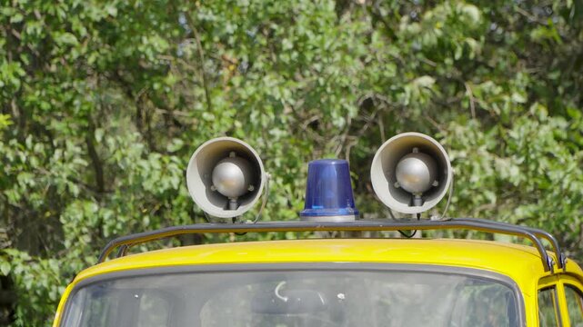 Two megaphones and a blue flashing emergency beacon light sit mounted on top of a yellow car roof against a lush green tree background.