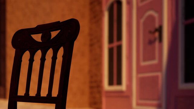 A wooden chair stands in front of a closed door inside a room. The light from outside creates shadows and highlights on the walls as evening approaches.