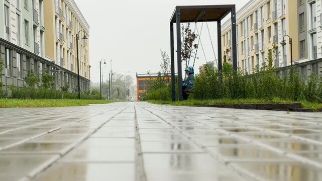 A young person sits quietly on a swing along a wet paved walkway between residential buildings. Rain falls gently in this courtyard. This dreary scene evokes a lonely and melancholic mood.