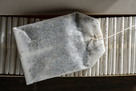 Single paper tea bag on top of organized row in box. Closeup top view of classic white tea bag with string resting horizontally over neat row of packed tea bags in cardboard package