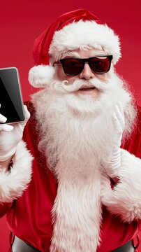 Santa Claus wears sunglasses and holds a smartphone. He smiles broadly, looking excited and amused. The vibrant red background adds festive energy. This video blends holiday cheer with modern tech