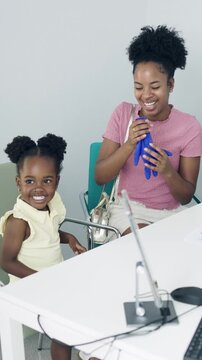 Playful African American mother inflating a medical glove like a balloon to entertain her laughing daughter while waiting in a clinic. Vertical