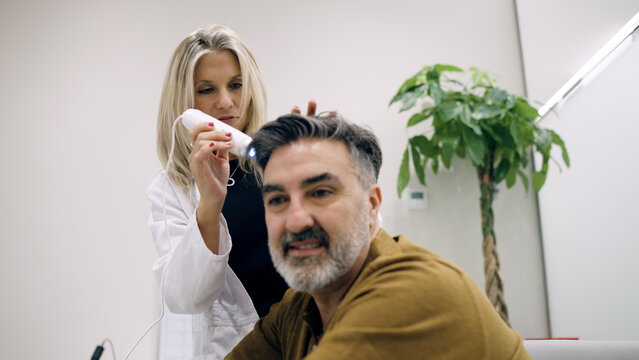 Female trichologist dermatologist using a dermatoscope inspecting male patient's scalp, analyzing hair loss problems in a clinic