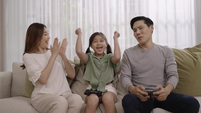 Happy family day concept. Asian father, mother, and daughter sitting using joystick and playing video game together having fun and cheerful on sofa in living room at home while child girl she winner. 