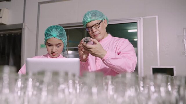 Two Asian quality control inspectors in uniform standing, using a laptop computer to analyze and inspect empty glass beverage bottles on moving conveyor belt in production line at industrial factory. 