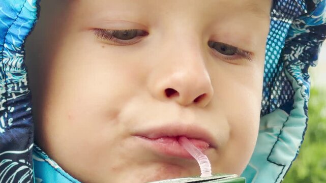 A young boy drinks juice through a straw outdoors. He wears a blue hooded jacket and feels calm and content while enjoying his sweet drink in a peaceful environment