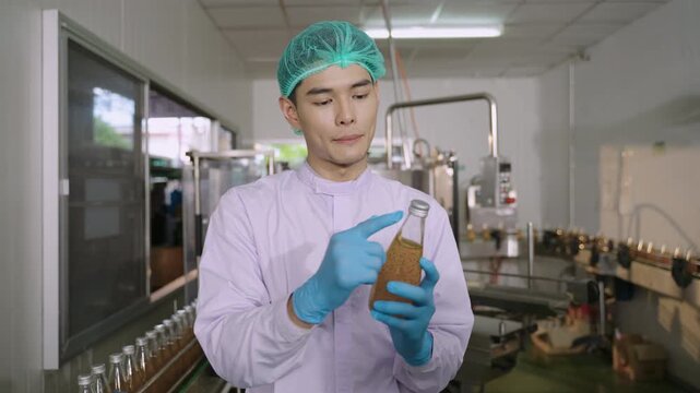 Professional Asian inspector in uniform of food and drink factory holding a juice beverage bottle promoting and advertising healthy nutritional benefits in production line at manufacturing industry.