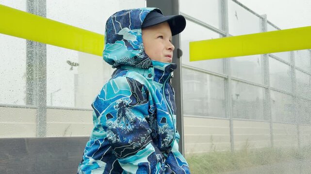 A young boy in a blue jacket sits on a bench waiting inside a rainy bus shelter. The quiet child looks thoughtful and calm during the storm. This melancholic environment feels lonely.