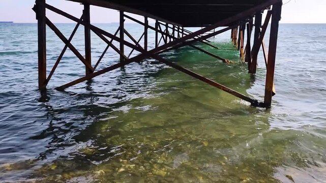 Cinematic View Underneath Old Metal Pier Structure Over Clear Blue Ocean Waves Rippling Water Surface Sea Beach Shore Line Pier Legs Steel Support Structure Daylight Water Surface Background