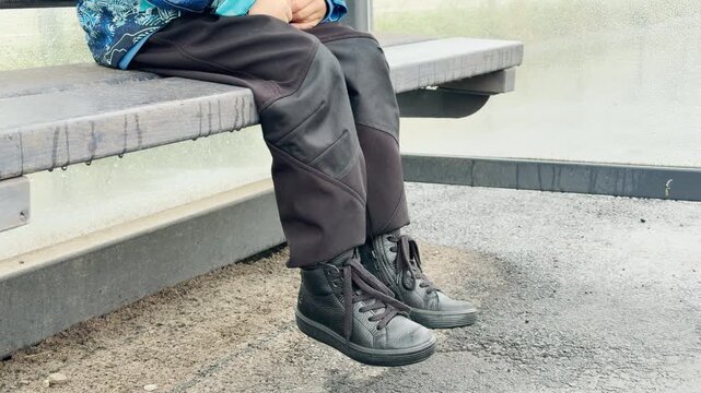 A child sits quietly on a wooden bench inside a bus shelter. The young person wears black pants and sneakers while waiting. This calm, melancholic scene feels peaceful yet lonely.