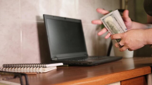 Close up of hands counting and fanning big stack of 100 US dollar bills near laptop on wooden table in home office workspace