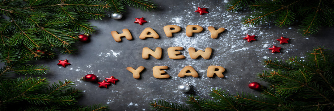 Happy new year text made of gingerbread cookies arranged on a dark grey surface with pine branches, red ornaments, and white flour dusting
