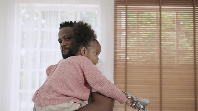 Happy African American father standing, holding, and embracing his cute little daughter with love and bonding in living room at home. A moment of love. A black dad carrying a child girl, affectionate.