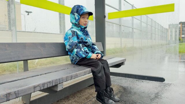 A young boy in a blue raincoat sits on a wooden bench. He waits inside a wet bus shelter during a rainy day. This dreary weather creates a lonely and melancholic mood.