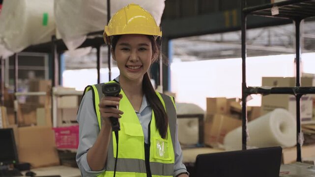 Portrait of happy Asian woman warehouse worker in uniform and wearing yellow safety helmet, standing, working, and holding barcode scanner while looking at camera in logistics distribution factory. 