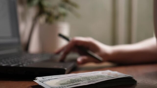 Close up focus on stack of 100 US dollar banknotes on desk with blurred person holding pen and using laptop touchpad in home office