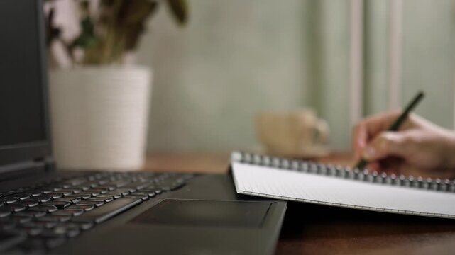 Close up side view of person writing notes with pencil in open notebook on wooden table next to laptop in home office