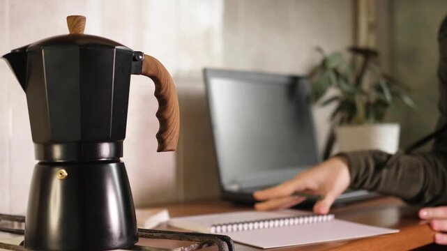 Person flipping notebook pages, thinking and writing notes with pencil near black moka pot coffee maker and laptop on table