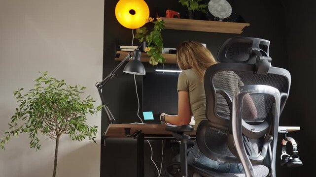 Woman works at a desk in a home office, sitting in an ergonomic chair with a computer and notebook.