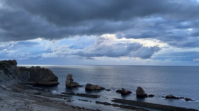 Layered storm clouds fill the twilight sky over a calm Atlantic Ocean, creating striking shades of grey above the dark rocky coast of Cantabria, Spain.