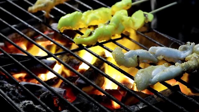 A vendor's hand turns skewered squid roe and meat marinated in golden turmeric over blazing charcoal, producing dramatic flame flare and smoke at a Thai night market stall.