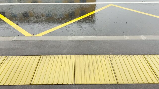 Yellow tactile paving sits on a wet sidewalk as rain falls on the asphalt road. The dreary weather creates a lonely and melancholic mood in the urban street.